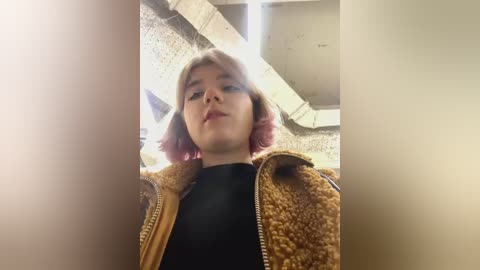 Media: Video of a young woman with short, pink hair, wearing a black shirt and a brown, fuzzy jacket, standing in an indoor, concrete corridor with fluorescent lighting.