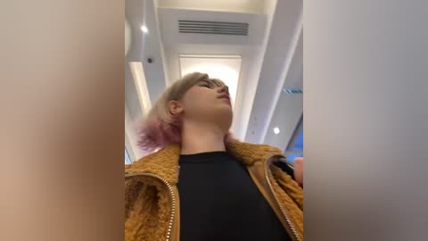 Media: A video of a young woman with short, light brown hair and pink highlights, wearing a black shirt and a brown vest, standing inside an airplane aisle. The background features white ceiling panels and a modern interior design.