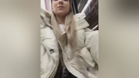 Media: Video of a young woman with light skin, long blonde hair, and a white puffy jacket, standing in a dimly lit, narrow space with metal walls.