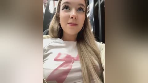 Media: Video of a young Caucasian woman with long blonde hair, wearing a white T-shirt with a large pink bow graphic, sitting in a dimly lit, blurred background.