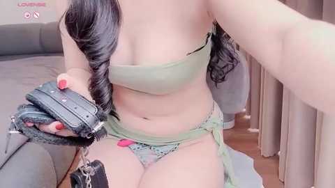 Media: A video of a woman with fair skin, long dark hair in a braid, wearing green lingerie and a black harness, sitting on a bed with beige curtains in the background.