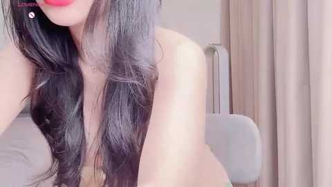 Media: A video of a topless woman with long, wavy black hair, fair skin, and red lipstick, sitting on a white chair in a softly lit room with beige curtains.