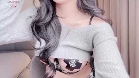 Media: Video of a light-skinned woman with long, wavy dark hair, wearing a light gray, off-the-shoulder sweater and a black and white bra with \"XXL\" printed on it, in a cozy indoor setting.