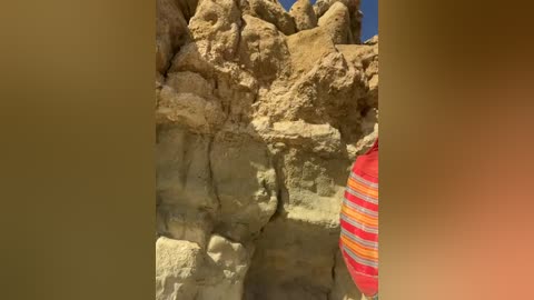 Media: A video capturing a rugged, beige rock formation with a textured surface under a clear blue sky. The right side features a partially visible person wearing a red and yellow striped shirt.