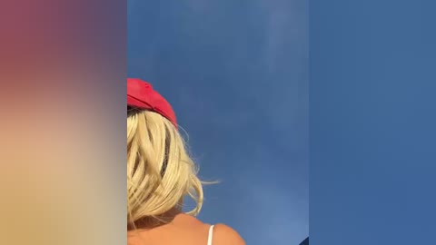 Media: Video of a blonde woman with shoulder-length hair, wearing a red hat, facing away, against a blue sky.