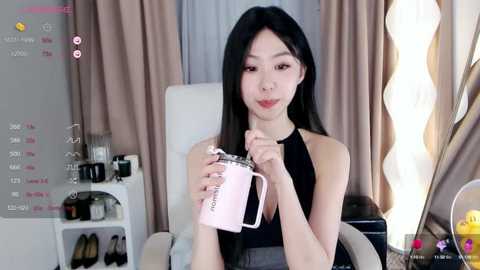 Media: Video of an East Asian woman with long black hair, wearing a black halter top, holding a pink cup in a modern living room with beige curtains and a white chair.