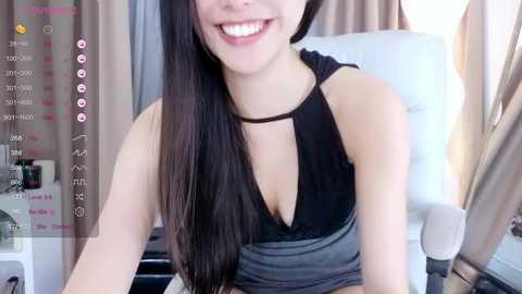 Media: Video of a smiling Asian woman with long black hair, wearing a black halter top, sitting on a white chair in a modern room with beige curtains.