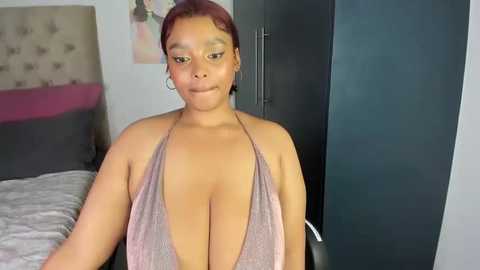 Media: Video of a plus-sized Black woman with medium-brown skin and auburn hair, wearing a revealing halter top that highlights her large breasts, sitting on a bed in a modern, minimalistic bedroom.