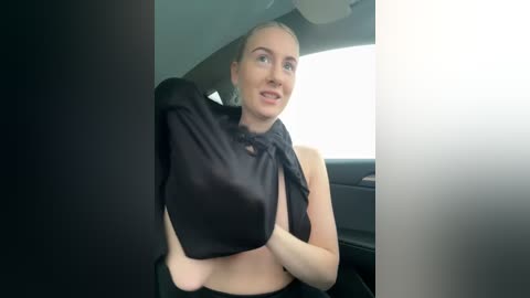 Media: Video of a fair-skinned woman with blonde hair, wearing a black shirt, adjusting it in a car, with a blurry, sunlit background.