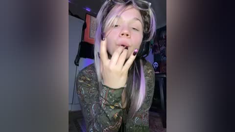 Media: A video of a young woman with pale skin, long blonde hair, and dark-rimmed glasses, wearing a green paisley shirt, sitting in a dimly lit room, holding her face with her fingers.