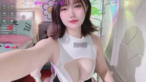 Media: Video of an Asian woman with straight black hair and fair skin, wearing a revealing white crop top that exposes ample cleavage. She sits in a cozy, colorful bedroom with playful decor.