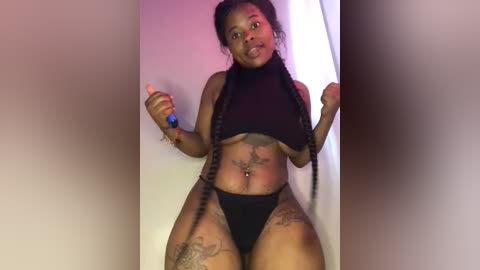 Media: Video of a dark-skinned woman with braids, wearing a black crop top and thong, holding a toy. She has tattoos on her thighs, arms, and chest.