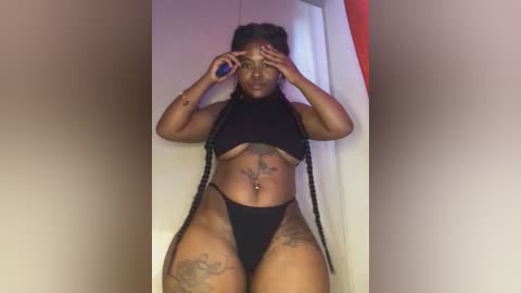 Media: Video of a dark-skinned woman with a voluptuous figure, wearing a black bralette and thong, showcasing tattoos on her thighs and belly, standing in a bathroom with a red curtain in the background.