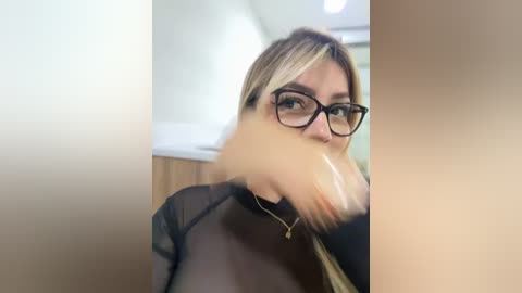 Media: Video of a blonde woman with glasses, wearing a sheer black top, eating a sandwich, in a modern, light-colored room.