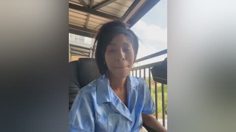 Media: Video of a woman with short black hair, wearing a light blue button-up shirt, seated on a dark cushioned chair on a balcony with metal railing.