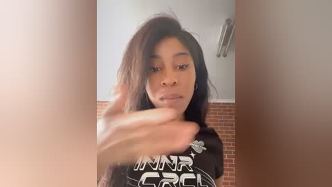 Media: Video of a young Black woman with long, straight dark hair, wearing a black t-shirt with white text. She has a neutral expression. Background includes a brick wall and white ceiling.