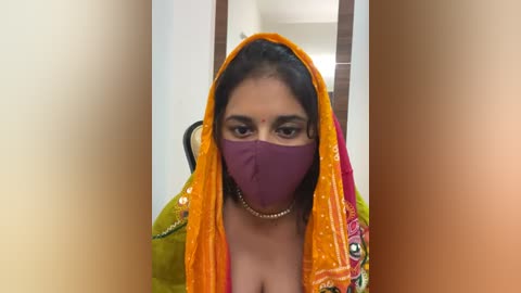 Media: Video of a South Asian woman with medium skin tone, wearing a bright orange sari, purple mask, and gold jewelry, seated indoors with a blurred background.