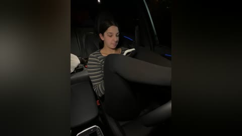 Media: Video of a young woman with medium-length dark hair, wearing a striped sweater and black leggings, sitting in a car with dim lighting, looking down at a phone.