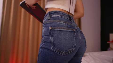 Media: Video of a woman's backside, wearing tight, dark blue jeans with a white top, holding a black book. Background includes beige curtains and a bed.