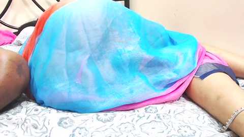 Media: Video of a person lying on a bed, wearing a loose, bright blue and pink patterned dress, with a black bra strap visible.