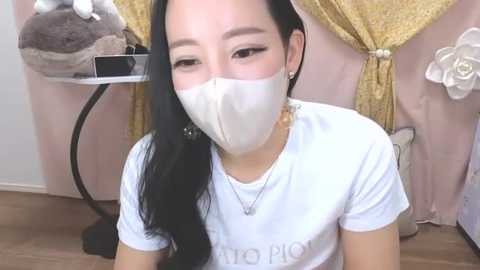 Media: Video of an Asian woman with long black hair, wearing a white mask, white t-shirt, and a gold necklace, sitting on a wooden floor, with a grey cat toy and pastel-colored wall decor in the background.