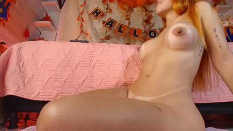 Media: Video of a nude, pale-skinned woman with long red hair, sitting on a pink quilted bedspread, with a Halloween-themed banner in the background.