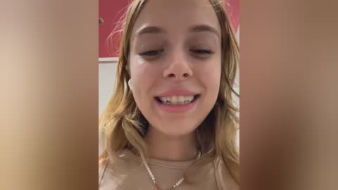 Media: Video of a young Caucasian woman with light skin, wavy blonde hair, and a friendly smile. She wears a beige top and white earbuds. Background shows a red wall and white ceiling.