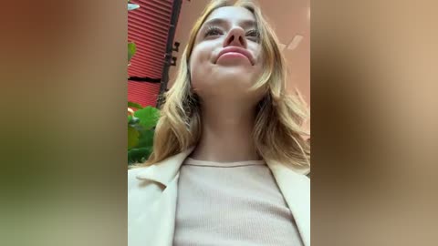 Media: Video of a smiling, light-skinned woman with blonde hair, wearing a beige blazer over a cream-colored top, standing indoors near a red window with green plants in the background.