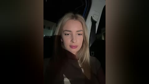 Media: Video of a young woman with straight, shoulder-length blonde hair, wearing a black coat and red lipstick, seated in a dimly lit car, looking contemplative.