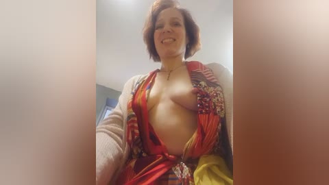 Media: A video of a fair-skinned, short-haired woman with a warm smile, wearing an open, colorful robe exposing her small breasts. Background shows a neutral-toned room with a window.