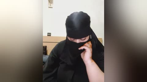 Media: Video of a person wearing a black niqab, standing in a dimly lit room with a beige wall and a small wooden table.