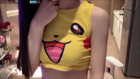 Media: Video of a slender, light-skinned woman in a sleeveless yellow crop top featuring Pikachu's face, wearing a white tank top underneath. She is seated on a white chair in a modern, beige-toned kitchen with stainless steel appliances and floral arrangements.