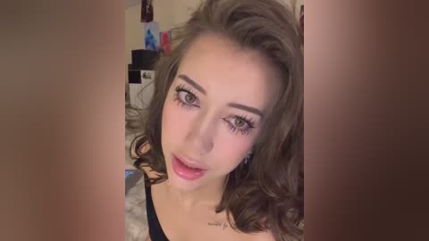 Media: Video of a young Caucasian woman with wavy, shoulder-length brown hair, wearing black clothing, and makeup featuring long eyelashes and pink lipstick, in a cozy indoor setting.