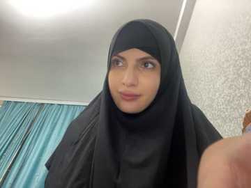 Media: Video of a young woman in a black hijab, with a soft expression, in a room with teal curtains and textured wallpaper.