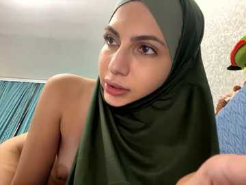 Media: A close-up video of a topless woman with light brown skin and medium-sized breasts, wearing a green hijab, in a room with a blue curtain and patterned wallpaper.