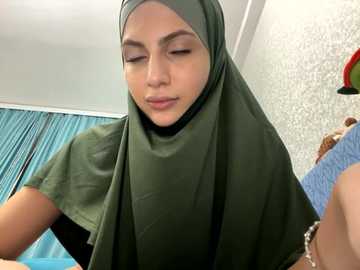 Media: Video of a young woman with a light brown complexion wearing a green hijab and a black shirt, looking down thoughtfully in a room with blue curtains and textured wallpaper.