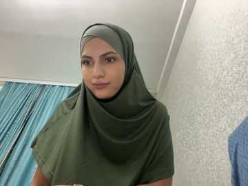 Media: Video of a young woman with light brown skin, wearing a green hijab, standing in a room with white walls and a blue curtain.