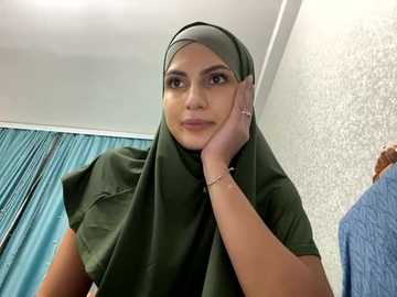 Media: Video of a light-skinned woman with medium-length dark hair, wearing a green hijab and a casual top, resting her chin on her hand, in a room with teal curtains and textured wallpaper.