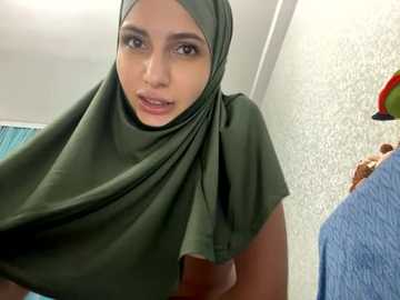 Media: Video of a young woman with light brown skin and dark eyes, wearing a green hijab and a light blue top. Background includes a textured wall and a blue towel hanging.