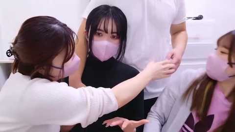 Media: Video of four Asian women in white lab coats and pink masks, assisting a seated Asian woman with a black headscarf, in a bright, sterile laboratory setting.