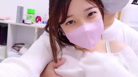 Media: Video of an East Asian woman with light skin, wearing a white mask, and a white shirt, taking a selfie in a white room with a white cabinet and various toiletries.