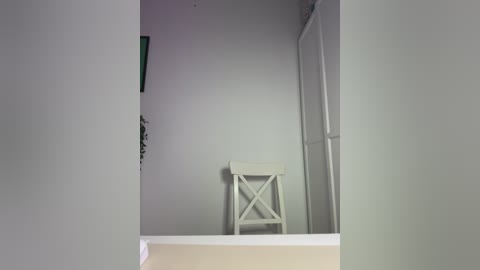 Media: Video of a minimalist, sparsely furnished room with a white wooden stool in the center against a plain, light gray wall. The image is taken from a low angle, looking up from the floor.