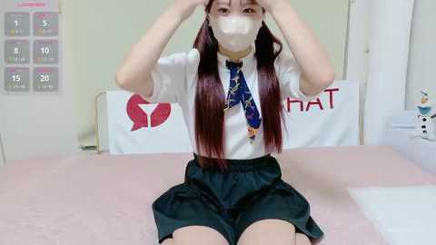 Media: Video of an Asian woman with long, straight, maroon hair, wearing a white blouse, black skirt, and mask, sitting on a pink bed, in a clean, simple bedroom with white walls and a poster.