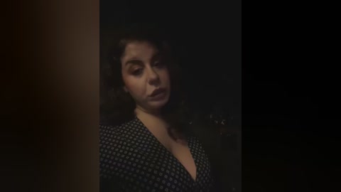 Media: A dimly-lit video featuring a woman with curly dark hair, wearing a black and white patterned dress, set against a dark background, conveying a somber, introspective mood.