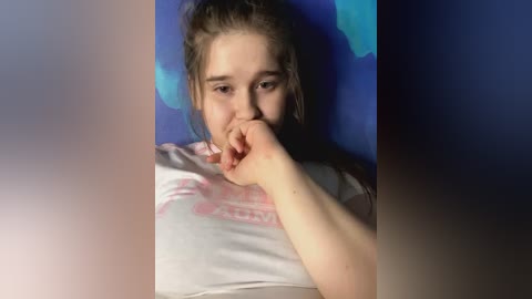 Media: Video of a young Caucasian girl with light skin and brown hair, wearing a white t-shirt, covering her mouth with her right hand. She has a serious expression. The background features a blue and white globe.