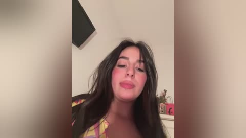 Media: Video of a young woman with long, dark hair, wearing a colorful plaid shirt, with flushed cheeks, standing in a room with a black shelf and small pink objects.