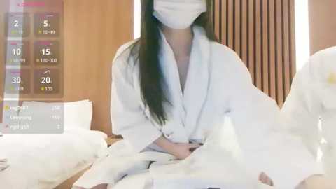 Media: Video of an Asian woman with long black hair, wearing a white robe, face mask, and glasses, lying in a bed with white sheets and wooden headboard, with a weather forecast overlay.