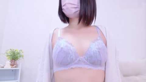 Media: Video of an East Asian woman with short, dark hair, wearing a white face mask, a white lace bra, and an open white robe, standing against a plain white background.