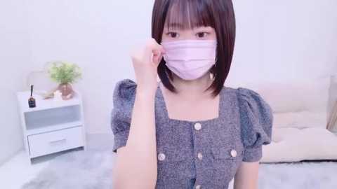 Media: A video of an Asian woman with straight, shoulder-length dark hair and a pink face mask, adjusting it in a minimalist bedroom with white walls, a white nightstand, and a beige cushioned chair.