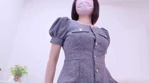 Media: A video of an Asian woman with a short bob haircut, wearing a gray textured button-up dress, a face mask, and sitting indoors.